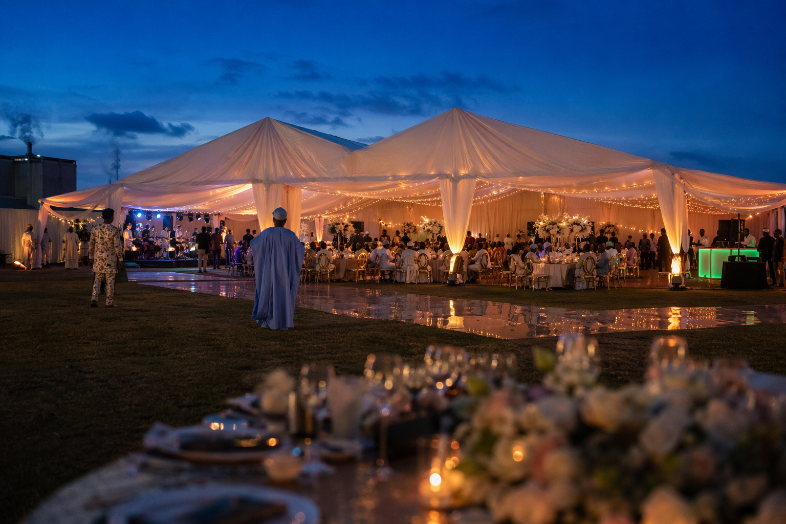 Outdoor Nigerian wedding reception at blue hour: white marquee draped with warm festoon lighting, guests in agbada seated at round tables with gold chairs, a reflective dance floor, deep cobalt sky overhead.